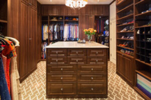 Turning a Spare Room Into a Closet Room-size walk-in closet with marble-topped dark wood chest of drawers in the center, a chandelier above, and matching wood shelves and sliding doors on either side